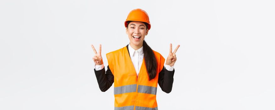 Happy upbeat asian female asian architect in safety helmet and reflective jacket, showing peace sign and smiling, assured in victory of her company, winning tender on construction works