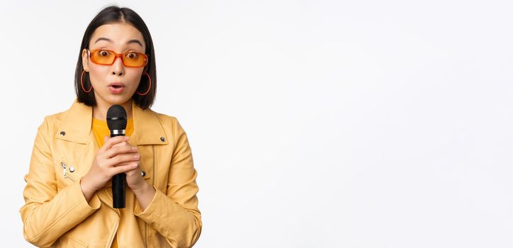 Portrait of beautiful asian woman in sunglasses, stylish girl singing, giving speech with microphone, holding mic and smiling, standing over white background
