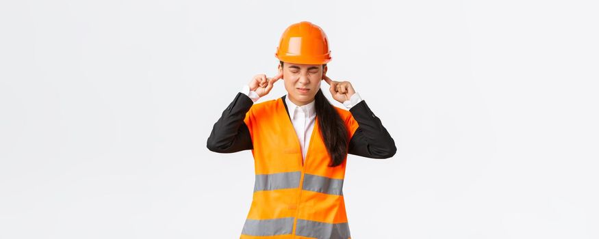 Asian female company manager feeling discomfort, chief engineer shut ears and grimacing from annoying noise, inspect work at construction zone, awful sound of repairment, wear safety helmet
