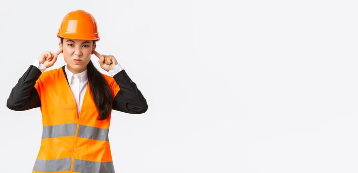 Bothered and displeased asian female chief engineer shut ears and grimacing from awful loud noise at construction area, wearing safety helmet, complaining on bothering sound, white background