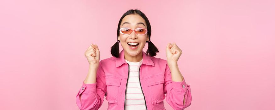 Portrait of excited japanese girl in sunglasses, celebrating, achieve goal, gasping amazed and smiling, standing over pink background