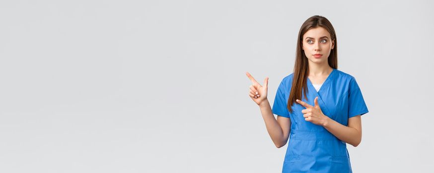 Healthcare workers, prevent virus, insurance and medicine concept. Dreamy cute nurse or doctor in blue scrubs, looking and pointing upper left corner, reading banner info, grey background.