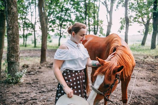 photo of a young smiling blonde, in a white blouse and skirt, with a horse, in a summer forest