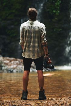 I want to capture photos of everything. Shot of a young man staring at a waterfall with his back turned and holding his camera.