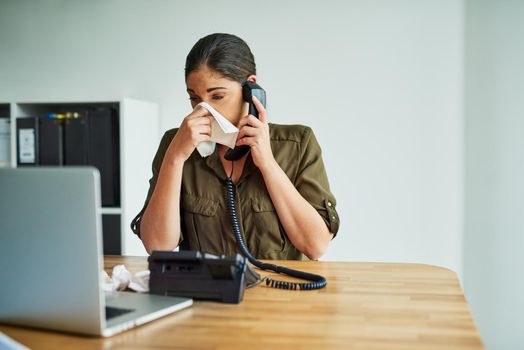 Its an unbearable condition to work in. Shot of a young businesswoman blowing her nose while speaking on a phone in an office.