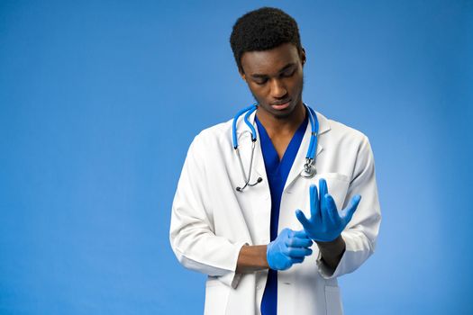 Young African American doctor putting on surgical gloves on blue background