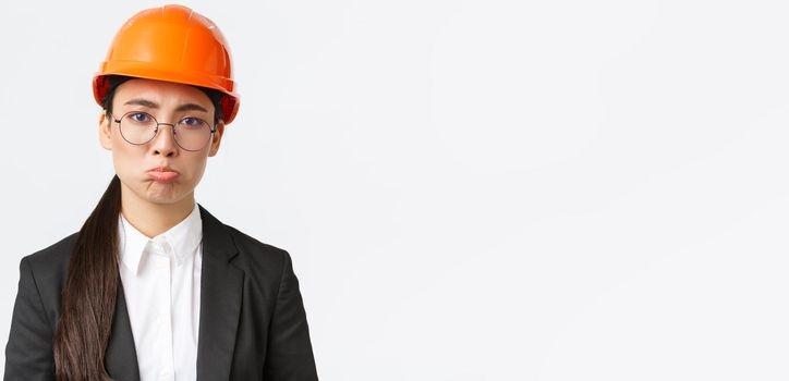 Close-up of upset and gloomy asian female construction manager facing failure, bad results on factory, engineer standing in safety helmet and business suit pouting sad, standing white background