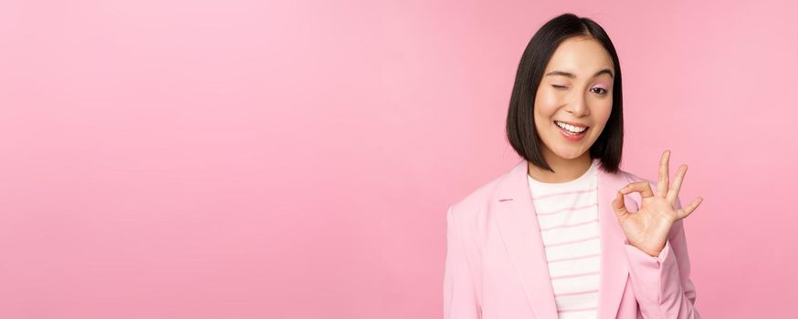 Excellent work, well done gesture. Smiling asian businesswoman showing okay, ok sign, praise good work, recommending company, looking as confident professional, pink background