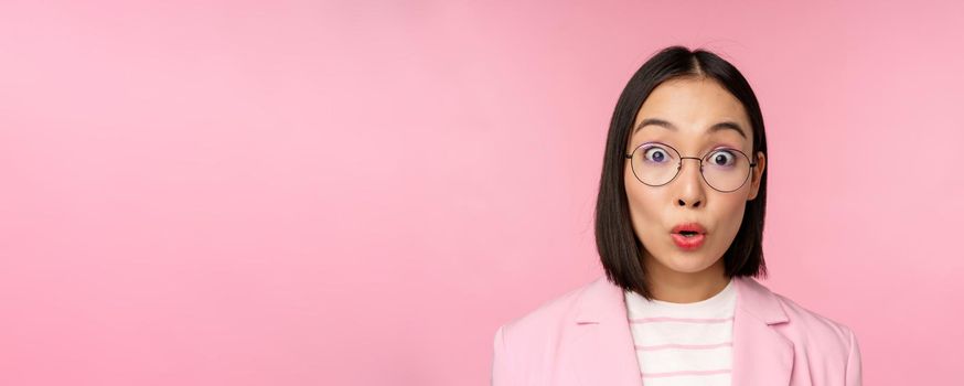 Close up portrait of asian businesswoman in glasses looking surprised at camera, amazed reaction, standing in suit over pink background