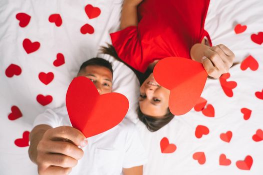 Top view of young attractive couple in love lying on the bed and showing red paper hearts. Happy valentine's day concept
