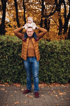Professional portrait of attractive young man embracing his little baby girl in plaid warm overall while standing in beautiful bright tree with red leaves. They are smiling at camera surrounded by vivid foliage in autumnal park.