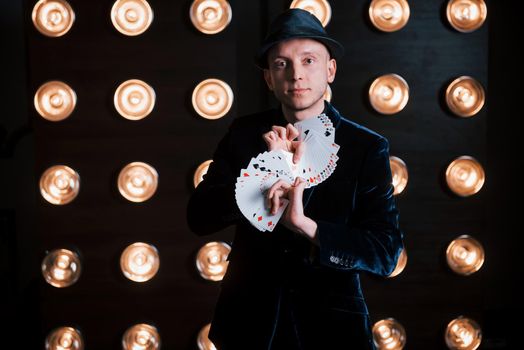 Conception of illusionist. Magician in black suit and with playing cards standing in the room with special lighting at backstage