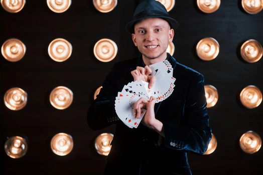 Your turn to pick. Magician in black suit and with playing cards standing in the room with special lighting at backstage