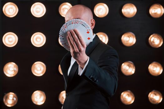 Focused photo on playing cards. Magician in black suit standing in the room with special lighting at backstage