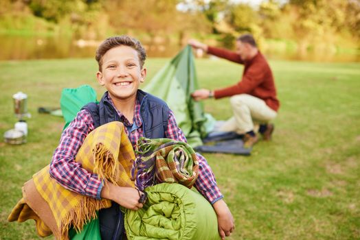 This camping trip is going to be awesome. Portrait of a young boy holding camping gear with his father in the background.