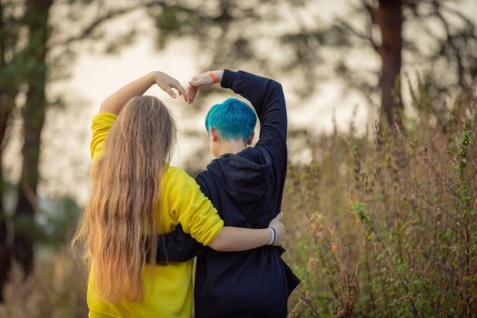 Two girlfriends are walking through the forest, raising their hands above their heads in the form of a heart. Girl with blue hair