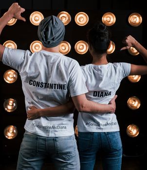 Shows thumbs on themselves. Back view of couple in shirts with their names on it standing next to studio lights