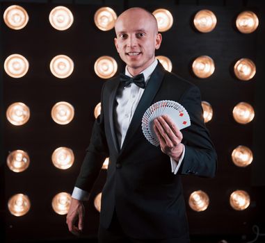 Smiling while showing deck of cards. Magician in black suit standing in the room with special lighting at backstage