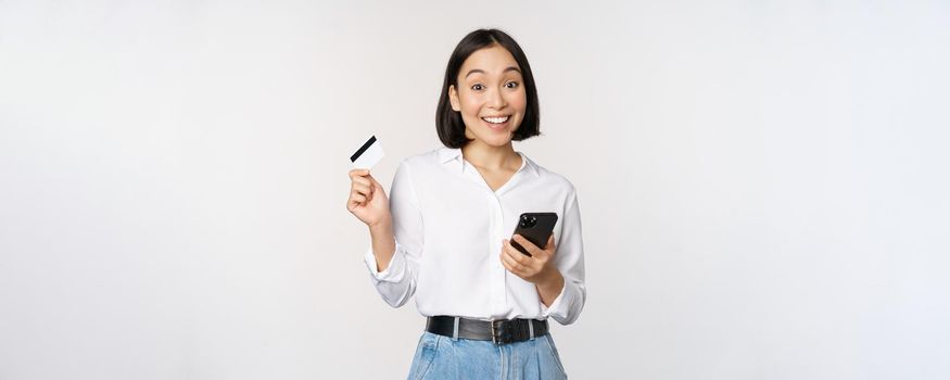 Online shopping concept. Image of young asian modern woman holding credit card and smartphone, buying with smartphone app, paying contactless, standing over white background