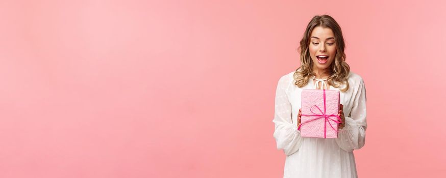 Holidays, celebration and women concept. Portrait of surprised charming young blond girl receive surprise gift, holding present in pink box look at it amused, curious whats inside, studio background