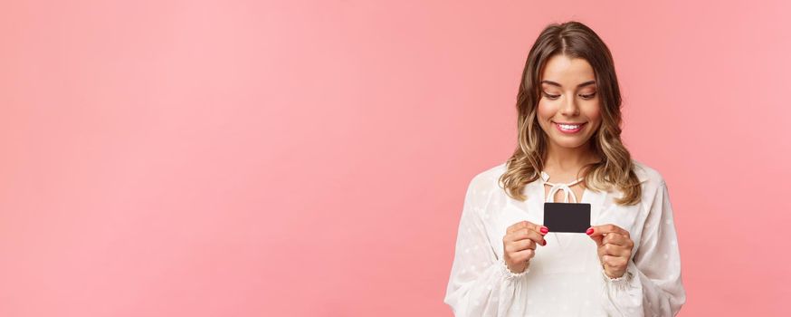 Close-up portrait of excited and amused blond girl in white dress, holding credit card and smiling thrilled, cant resist temptation to buy something, waste money online shopping, pink background