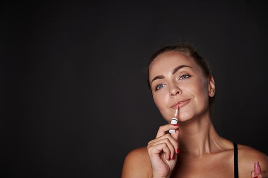 Beautiful woman applying lip gloss on her lips. Attractive blonde woman with moisturizing smoothing lip balm posing on black background with advertising copy space
