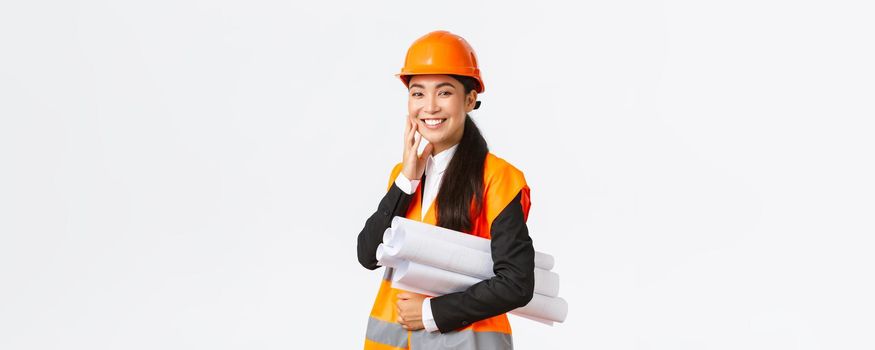 Smiling happy asian female architect, construction manager in safety helmet and jacket, carry blueprints of building project and looking delighted, finish plan in time, standing white background