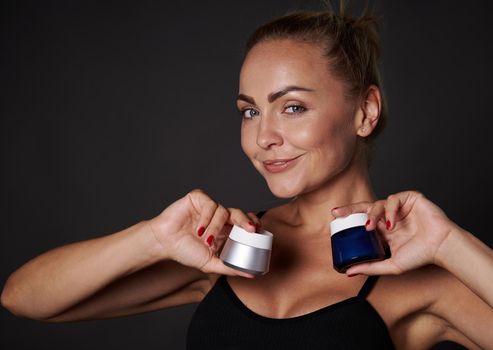 Beautiful middle aged blonde Caucasian woman holds jars with anti-aging anti-wrinkles smoothing facial cream , moisturizer, smiles at camera, isolated over black background with copy ad space