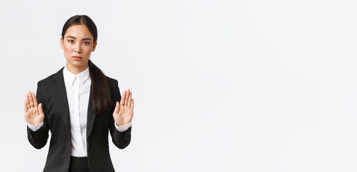 Serious-looking displeased asian female manager, businesswoman in black suit stop action, prohibit or restrict something, raising hands in forbid gesture, saying no or enough, white background