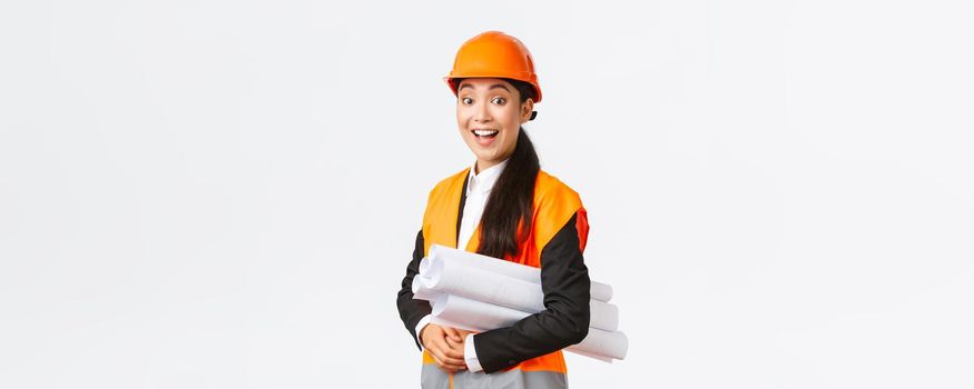 Amazed and excited smiling asian female architect, chief engineer in safety helmet and reflective jacket looking amused and happy, smiling impressed, holding blueprints of building area