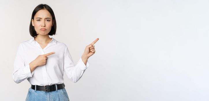 Sad and gloomy asian woman looking disappointed, complaining at banner or advertisement, pointing fingers right at promo and frowning upset, white background