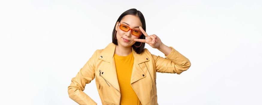 Stylish asian girl in sunglasses, looks cool and trendy, shows peace, v-sign gesture, stands over white background