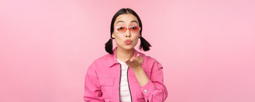 Stylish k-pop girl in sunglasses, asian woman blowing air kiss at camera, pucker lips, mwah gesture, standing over pink studio background