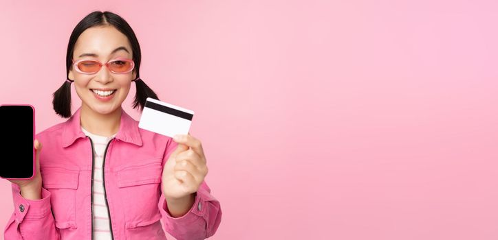 Image of smiling korean woman showing credit card and mobile phone screen, smartphone application interface, paying online, shopping contactless, standing over pink background