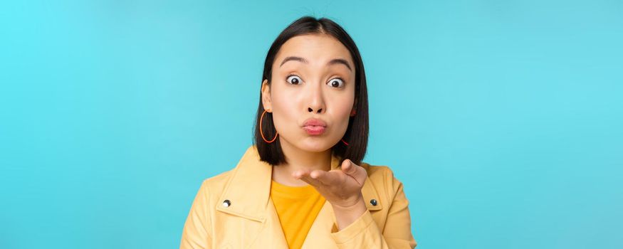 Close up portrait of funny asian girl sending air kiss, blowing at camera with popped eyes, standing over blue background
