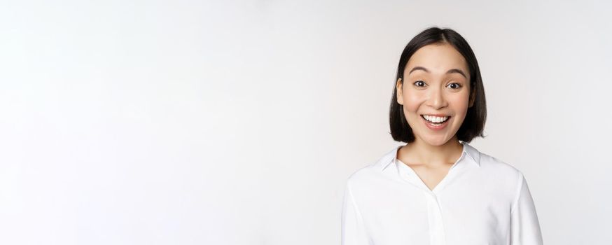 Close up portrait of young asian female model looking amazed at camera, smiling white teeth, standing against white background