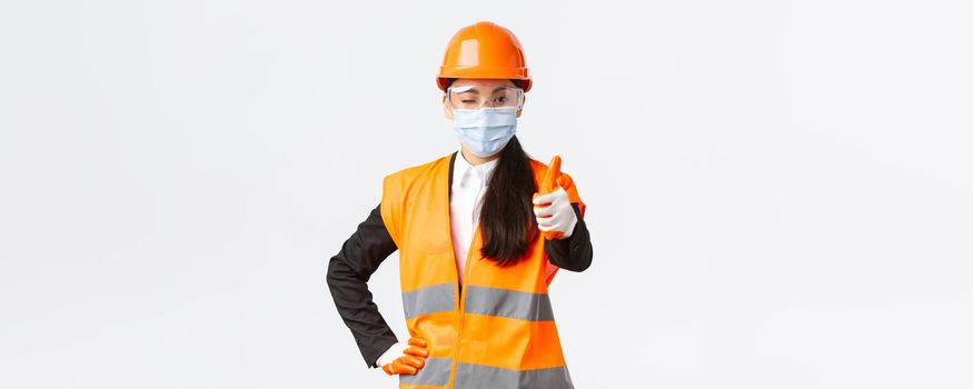 Covid-19 safety protocol at enterpise, construction and preventing virus concept. Confident asian female engineer, architect in face mask and helmet showing thumb-up, guarantee quality of building