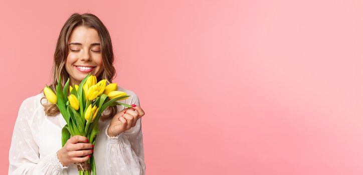 Spring, happiness and celebration concept. Close-up portrait of lovely, romantic blond girl sniffing smell of beautiful yellow tulips, close eyes and smiling happy, standing pink background.