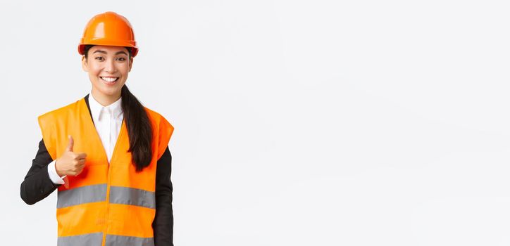 Confident satisfied asian female construction manager in safety helmet, reflective jacket, showing thumbs-up in approval, support and like work of employees, standing white background