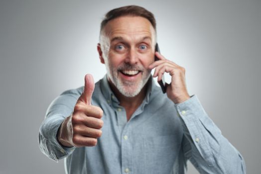 Weve been given the thumbs up to go ahead as planned. Studio portrait of a mature man showing thumbs up while talking on a cellphone against a grey background.