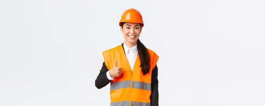 Confident satisfied asian female construction manager in safety helmet, reflective jacket, showing thumbs-up in approval, support and like work of employees, standing white background