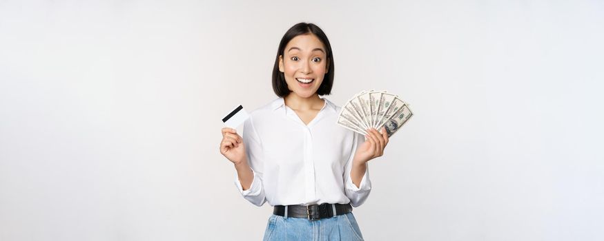 Portrait of enthusiastic asian woman holding money in cash and credit card, smiling amazed at camera, white background