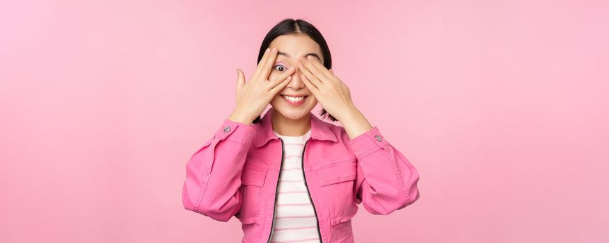 Portrait of asian girl peeks with excitement through fingers, covers eyes, seeing surprise, standing over pink background