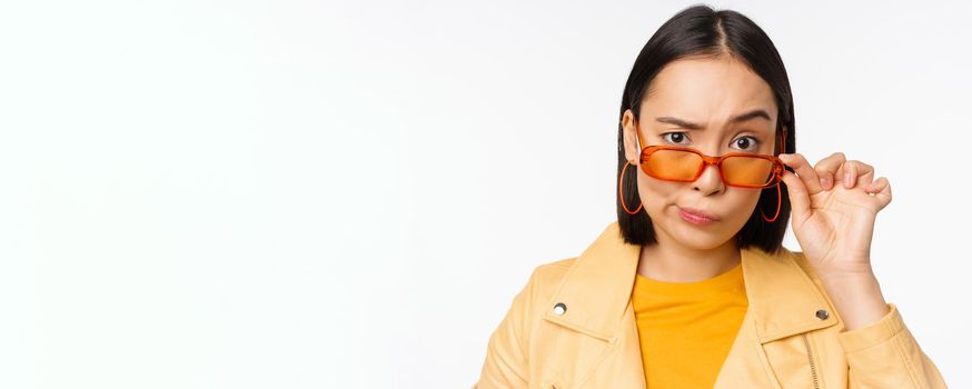 Image of young asian stylish woman, looking from under sunglasses with skeptical, disbelief face, express doubt, standing over white background