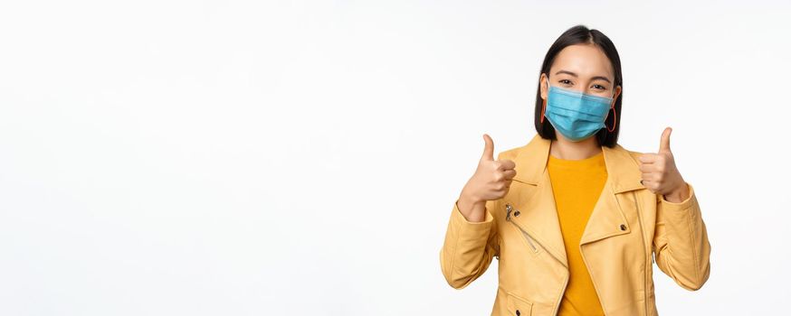 Smiling asian woman in medical face mask showing thumbs up, support wearing protective equipment from covid-19, coronavirus pandemic, white background