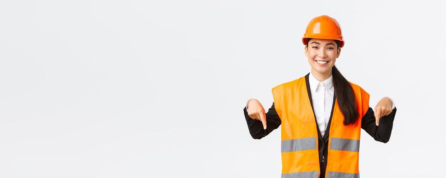 Smiling cheerful construction manager, asian female engineer in safety helmet and reflective clothing showing announcement. Woman project manager pointing down, standing white background