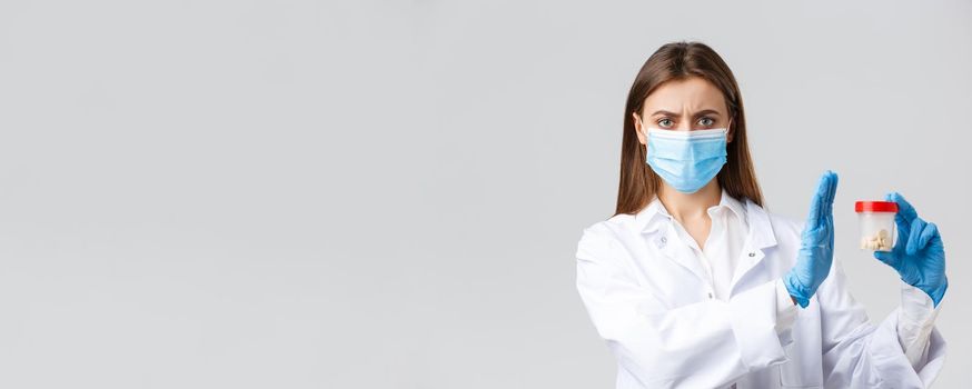 Covid-19, preventing virus, healthcare workers and quarantine concept. Concerned and displeased doctor in medical mask, gloves, reject, disapprove using medicine, showing bad pills.