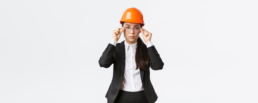 Lets get to business. Serious-looking confident female asian chief engineer inspect enterprise, wearing safety helmet and suit, put on glasses, standing white background, ready to work