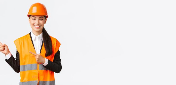 Building, construction and industrial concept. Smiling asian female architect in safety helmet, reflective clothing pointing finger upper left corner, showing project on workplace, white background
