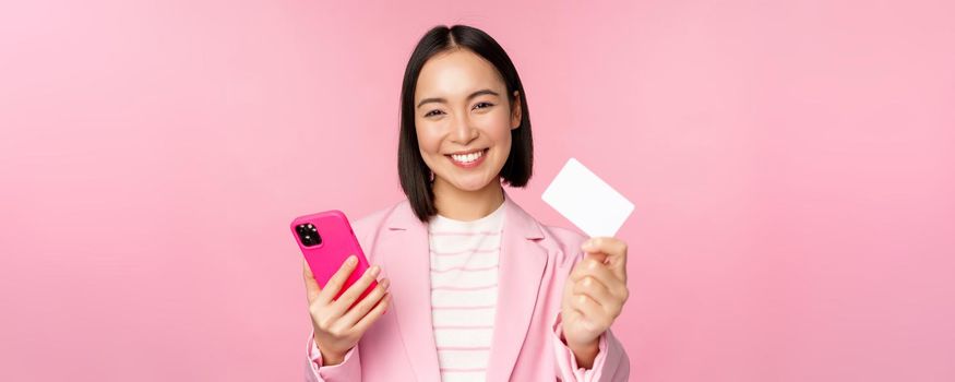 Image of smiling happy asian businesswoman showing credit card, paying online on smartphone application, order with mobile phon, standing against pink background